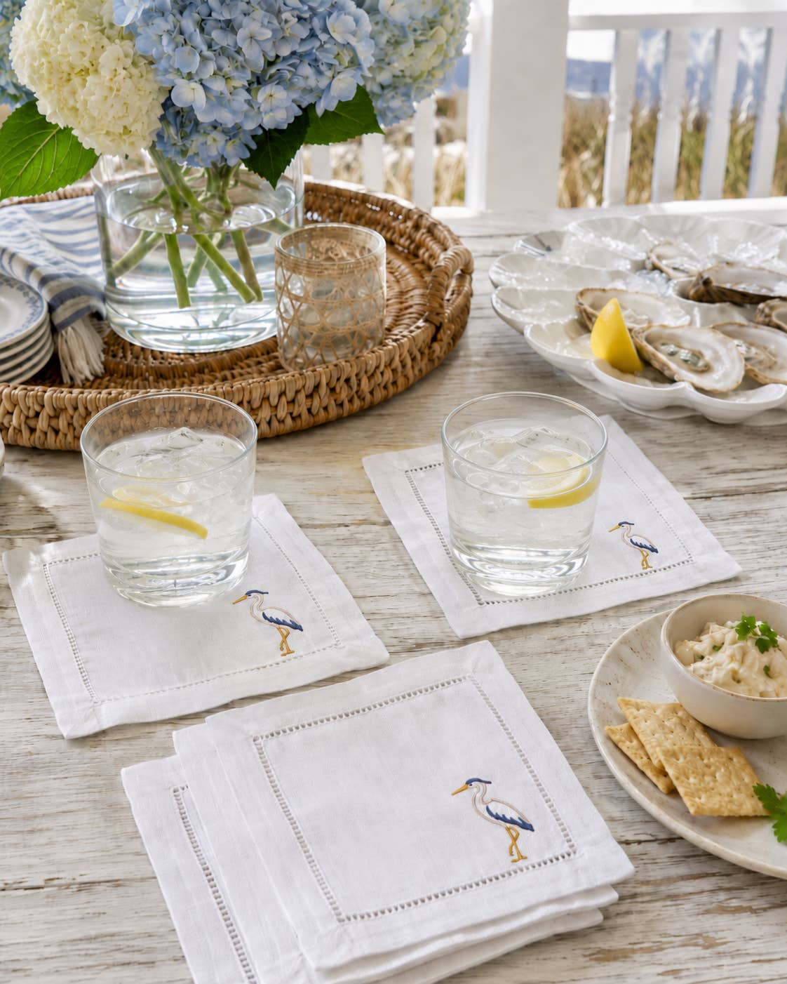 White embroidered linen napkins with a blue and yellow heron design, styled on a rustic outdoor table with glasses of water, oysters, crackers, and a floral centerpiece.