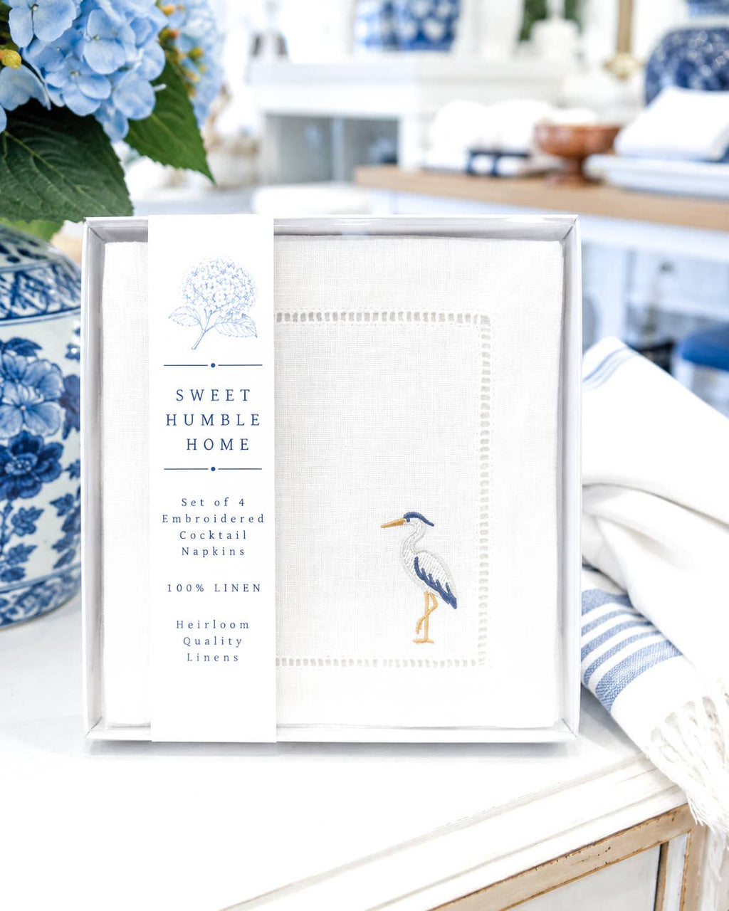A boxed set of four embroidered cocktail napkins from Sweet Humble Home, displayed on a white table with blue-and-white ceramic decor in the background. The napkins are white linen with blue, yellow, and orange embroidery details in a blue heron design.