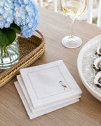 A stack of white linen napkins with a stitched border and a small heron embroidery on the front, styled on a wooden table with blue hydrangeas, a glass of white wine, and oysters on ice.