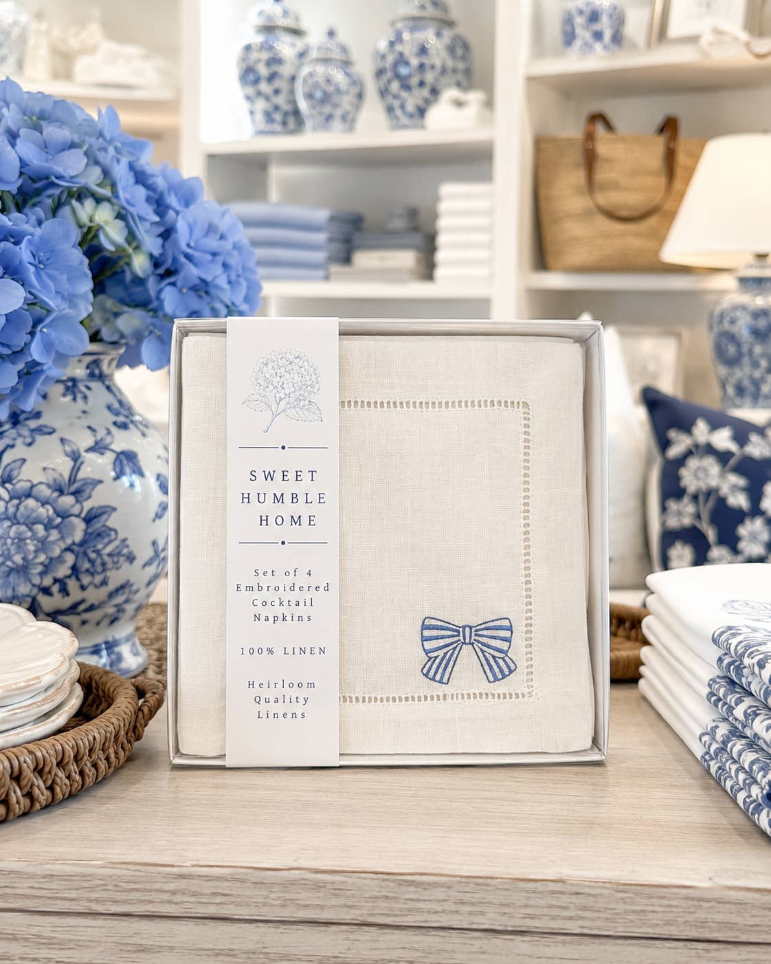 A boxed set of napkins from Sweet Humble Home displayed on a wooden table in a styled setting. The set includes a square linen napkin with hemstitch  border and bow details.
