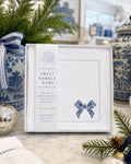 A boxed set of embroidered cocktail napkins from Sweet Humble Home, displayed on a marble surface with holiday greenery and blue-and-white ceramic decor in the background. The napkins are white with blue embroidery, including a bow motif, and the packaging includes a vertical label with the brand name.
