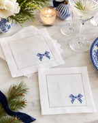 Two white linen napkins with blue embroidered bow details and stitched borders, arranged on a marble table with holiday decor, glassware, and a candle.
