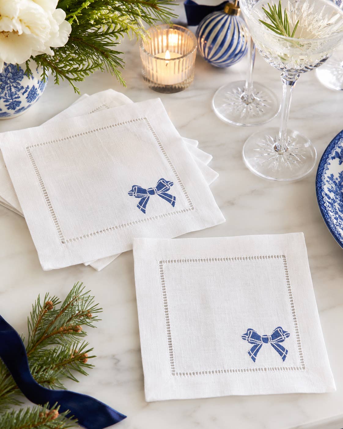 Two white linen napkins with blue embroidered bow details and stitched borders, arranged on a marble table with holiday decor, glassware, and a candle.