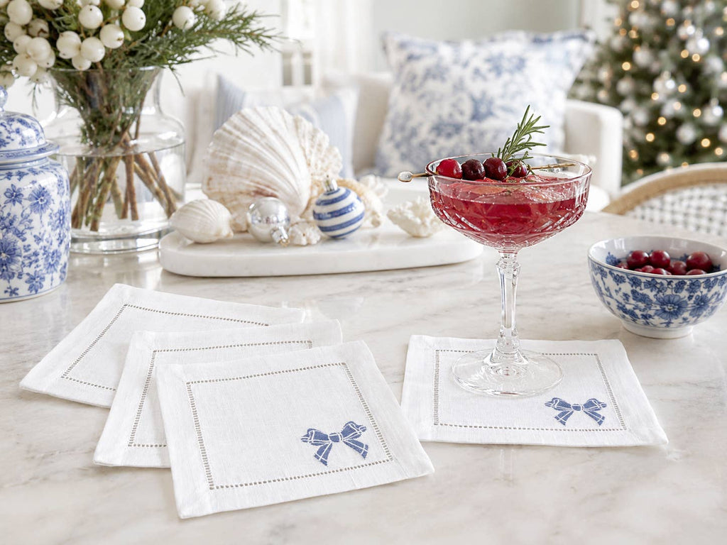 White linen napkins with blue embroidered bows and hemstitch borders, styled on a marble table in a holiday-themed setting with a blue-and-white patterned bowl, a cocktail glass, and seashells.