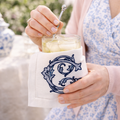 Person holding a glass with a drink and a decorative napkin with a monogram in a vintage style. The woman is in a pink cardigan and a light blue and white chinoiserie dress.