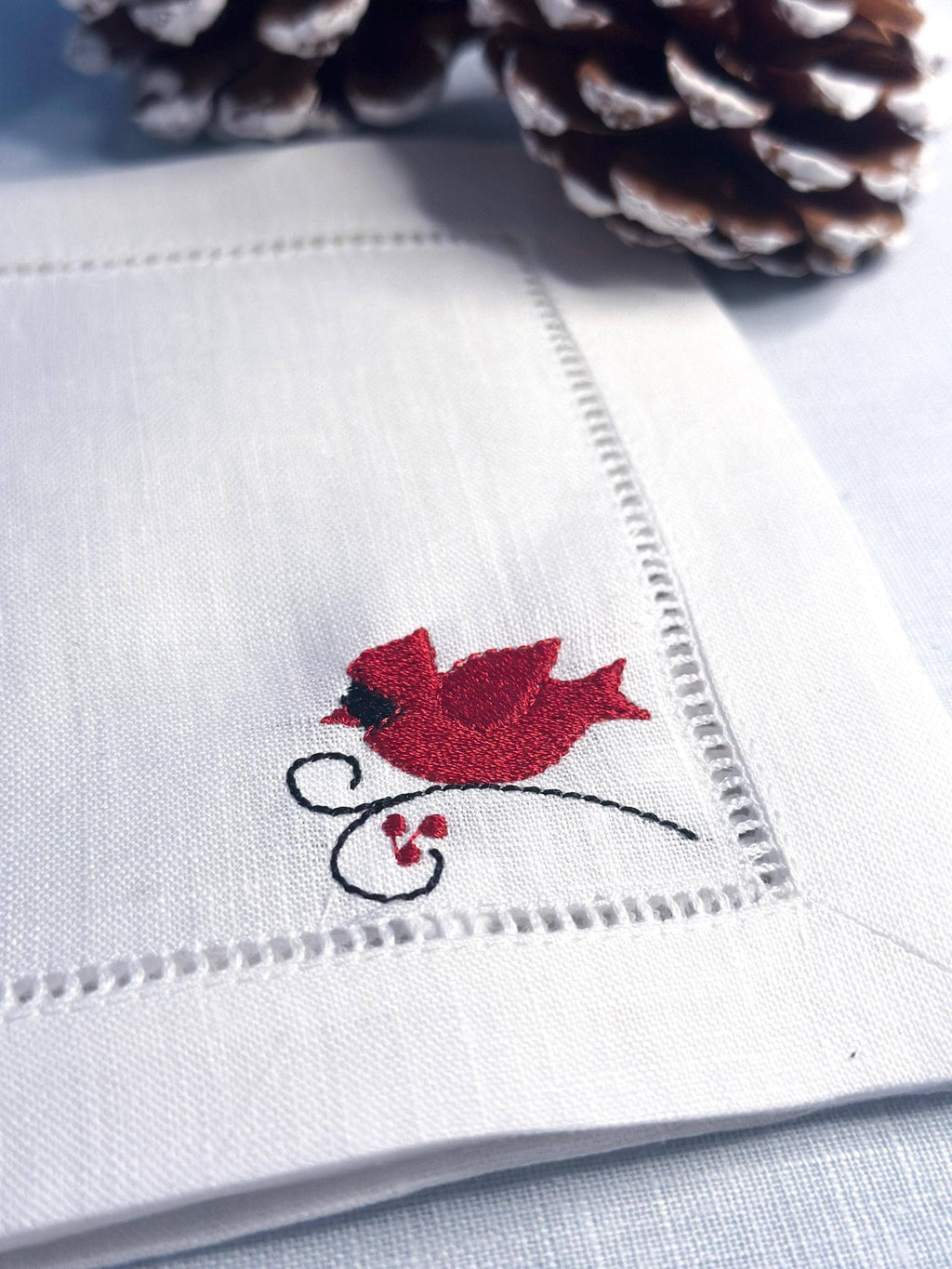 A close-up of a white embroidered napkin with a red cardinal bird and small red berries on one corner, styled with pinecones on a light blue surface.