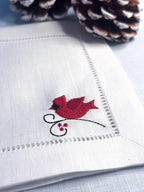 A close-up of a white embroidered napkin with a red cardinal bird and small red berries on one corner, styled with pinecones on a light blue surface.