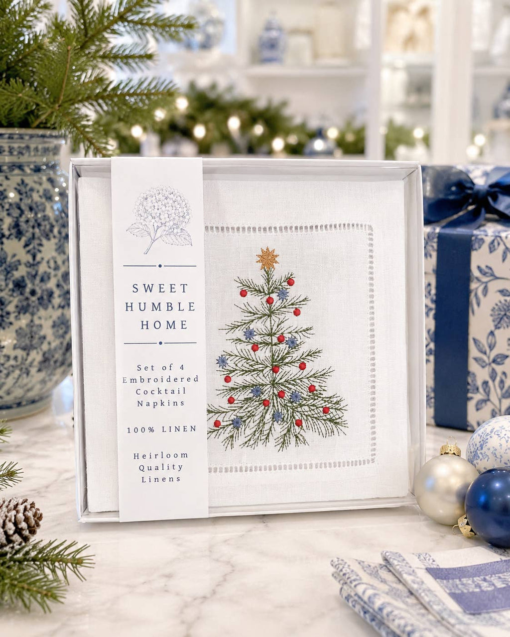 A boxed set of four embroidered cocktail napkins from Sweet Humble Home, featuring a white linen napkin with an embroidered green Christmas tree and red and blue ornaments, displayed on a marble surface with holiday decorations.