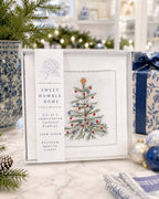 A boxed set of four embroidered cocktail napkins from Sweet Humble Home, featuring a white linen napkin with an embroidered green Christmas tree and red and blue ornaments, displayed on a marble surface with holiday decorations.
