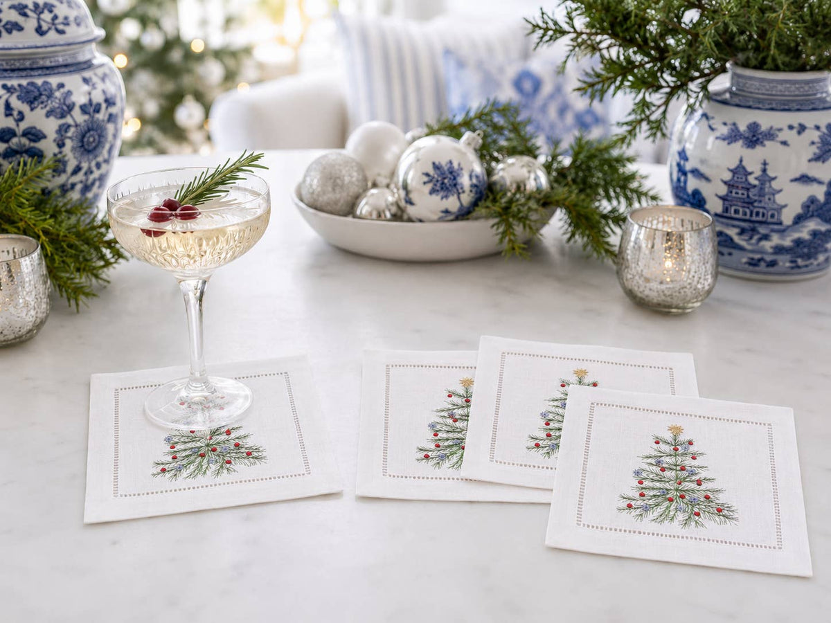 Four square white linen coasters with embroidered Christmas tree designs are arranged on a light marble table in a festive holiday setting, alongside a filled coupe glass, silver ornaments, greenery, and blue-and-white ceramic jars.