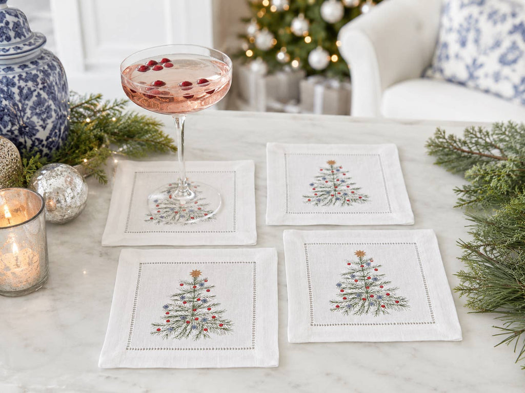 Four white linen coasters with embroidered Christmas tree designs arranged on a marble table, with a filled coupe glass, holiday greenery, ornaments, and a softly blurred festive interior in the background.