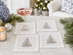 Four white linen coasters with embroidered Christmas tree designs arranged on a marble table, with a filled coupe glass, holiday greenery, ornaments, and a softly blurred festive interior in the background.