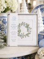 A boxed set of embroidered cocktail napkins from Sweet Humble Home, displayed on a tabletop with blue-and-white ceramic decor and holiday ornaments in the background. The napkin set includes a white linen napkin with a blue floral wreath embroidery and a vertical  label.