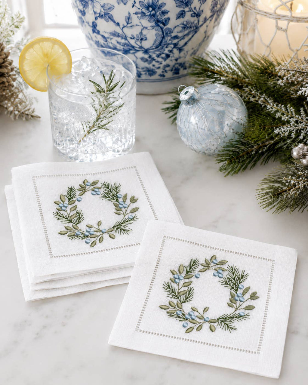 Two white embroidered napkins with a green and blue floral wreath design, styled on a marble surface with a glass of iced water, pine branches, and holiday ornaments in the background.