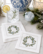 Two white embroidered napkins with a green and blue floral wreath design, styled on a marble surface with a glass of iced water, pine branches, and holiday ornaments in the background.