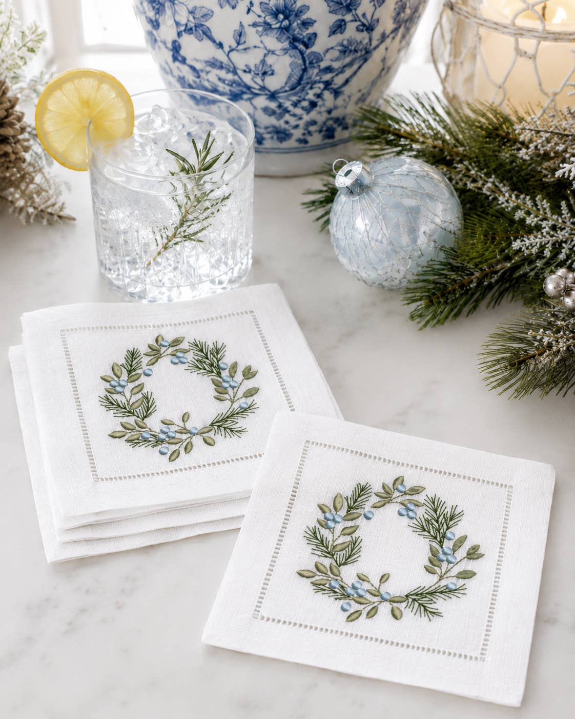 Two white embroidered napkins with a green and blue floral wreath design, styled on a marble surface with a glass of iced water, pine branches, and holiday ornaments in the background.