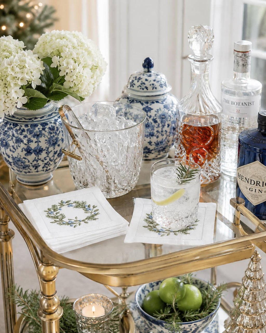 A styled bar cart scene with blue-and-white ceramic jars, a clear ice bucket, a glass of mixed drink garnished with rosemary and lime, embroidered cocktail napkins with floral wreath prints, and several liquor bottles in a bright indoor setting.
