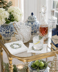 A styled bar cart scene with blue-and-white ceramic jars, a clear ice bucket, a glass of mixed drink garnished with rosemary and lime, embroidered cocktail napkins with floral wreath prints, and several liquor bottles in a bright indoor setting.