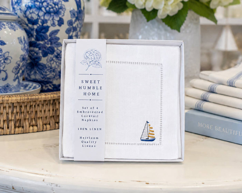 Close up of embroidered linen cocktail napkins with a sailboat in blue, white, and yellow above the hemstitch in Sweet Humble Home packaging on a table with a book, wicker basket, chinoiserie vases, and towels.