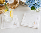 White linen cocktail napkins with sailboat embroidery on a table with a glass with rosemary and lemon.