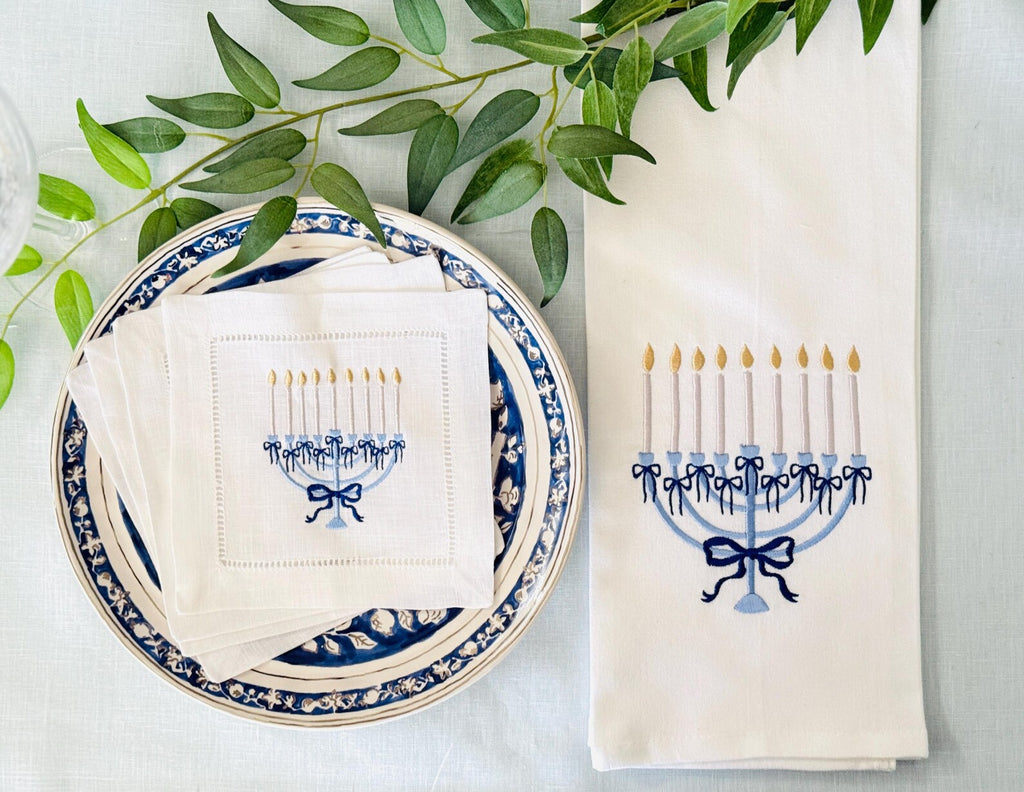 A flat-lay of white table linens featuring an embroidered Hanukkah menorah design in blue and gold. The linens are styled on a blue and white patterned plate with green eucalyptus leaves on a light blue tablecloth.
