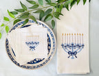 A flat-lay of white table linens featuring an embroidered Hanukkah menorah design in blue and gold. The linens are styled on a blue and white patterned plate with green eucalyptus leaves on a light blue tablecloth.