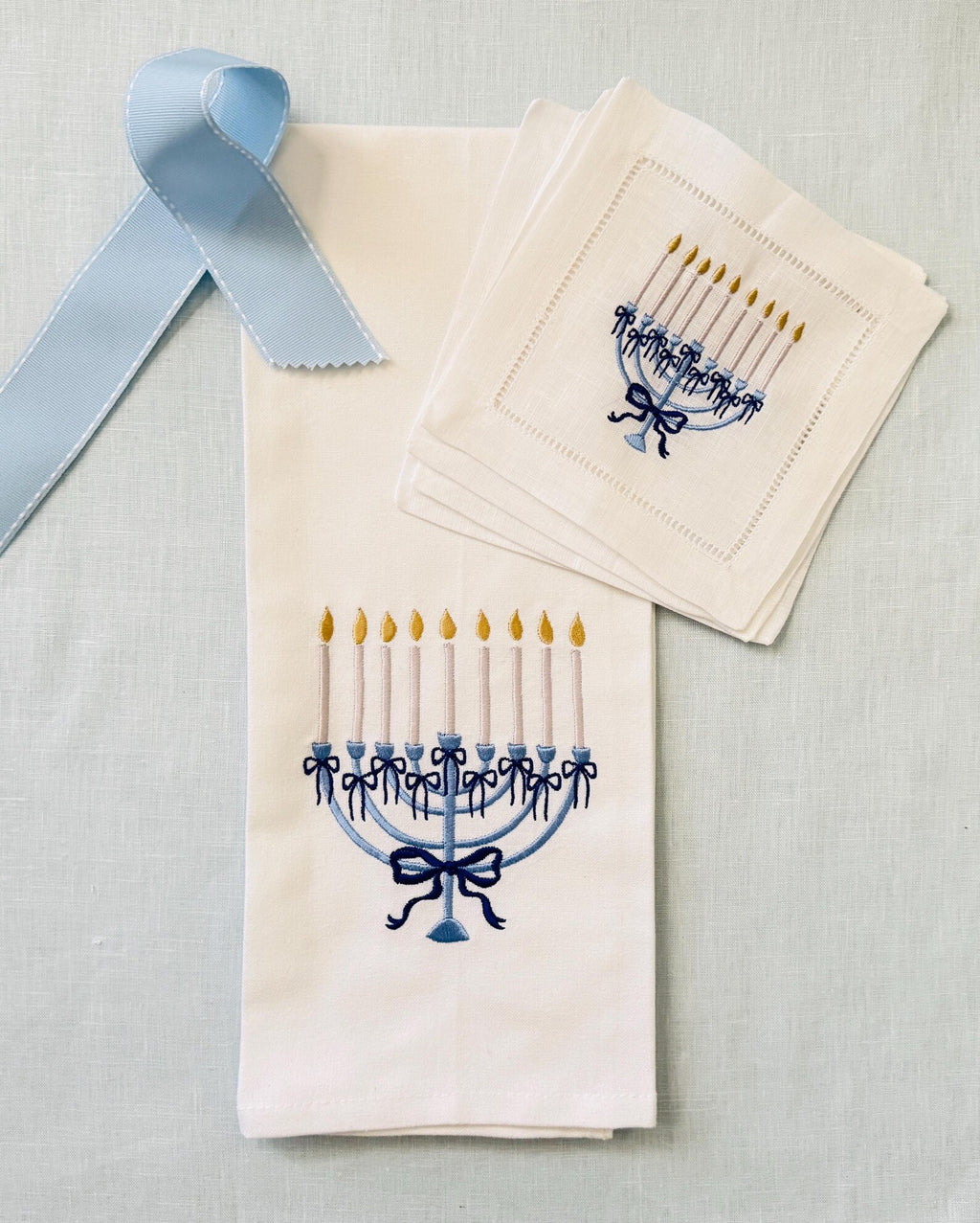A set of white linen cocktail napkins featuring embroidered blue menorahs with gold candle flames, and a white cotton kitchen towel embroidered with the same menorah arranged on a light blue linen textured background with a light blue ribbon.