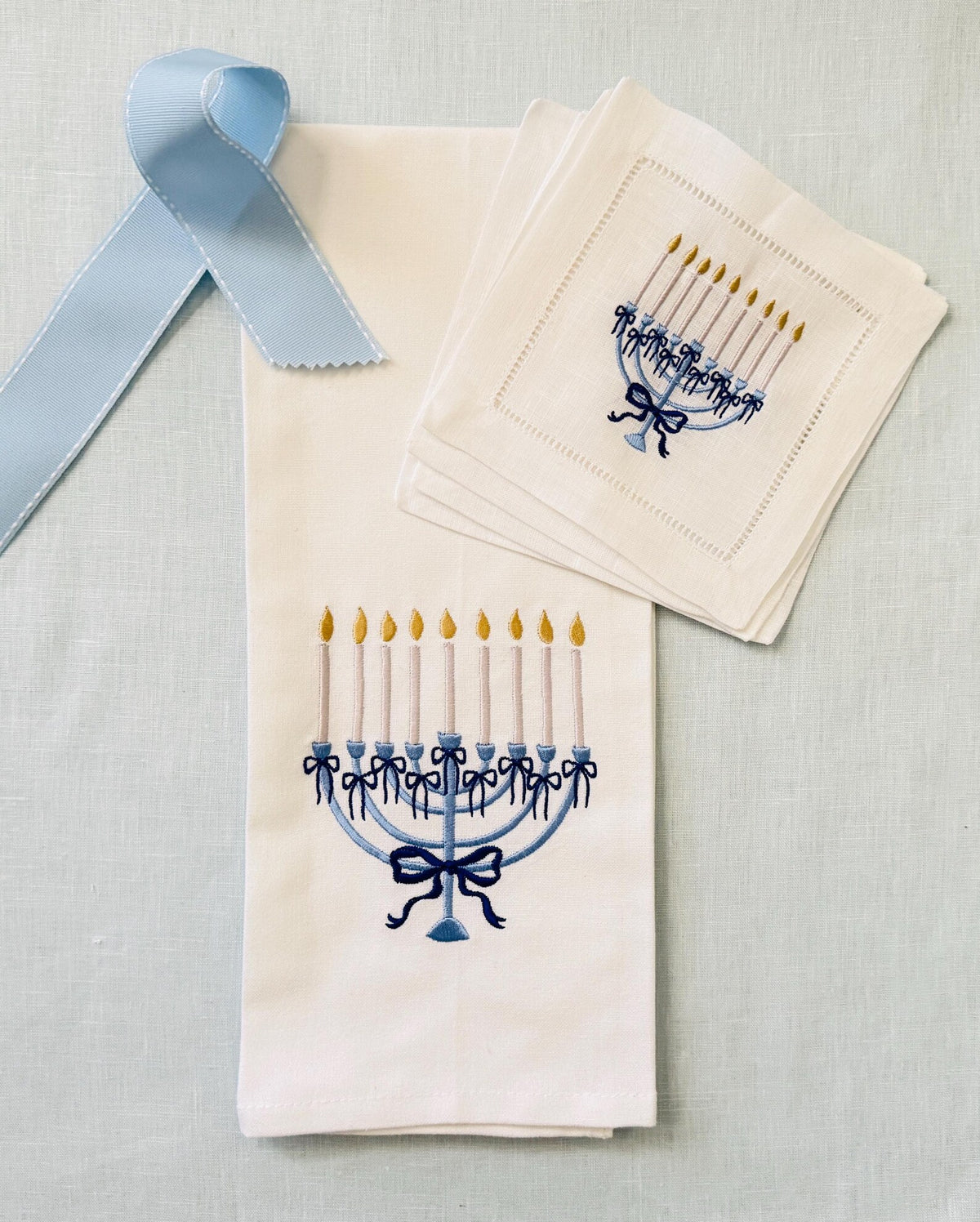 A set of white linen cocktail napkins featuring embroidered blue menorahs with gold candle flames, and a white cotton kitchen towel embroidered with the same menorah arranged on a light blue linen textured background with a light blue ribbon.