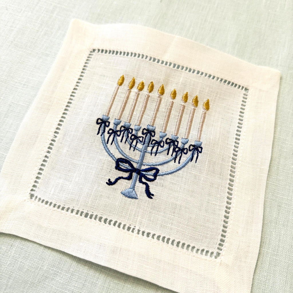 An embroidered square linen napkin featuring a blue menorah with gold candle flames and a navy bow, hemstitch border on a light blue textured linen background.
