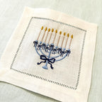 An embroidered square linen napkin featuring a blue menorah with gold candle flames and a navy bow, hemstitch border on a light blue textured linen background.