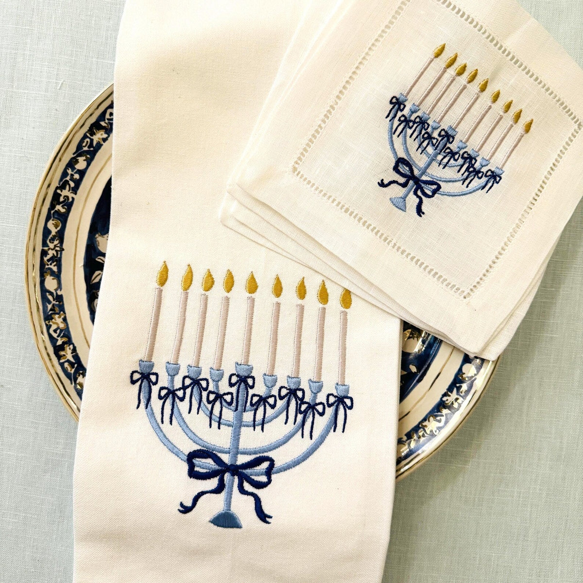 A set of white linen table linens, including a kitchen towel and 4 cocktail napkins, both featuring embroidered blue menorahs with yellow candle flames, styled on a patterned ceramic plate.