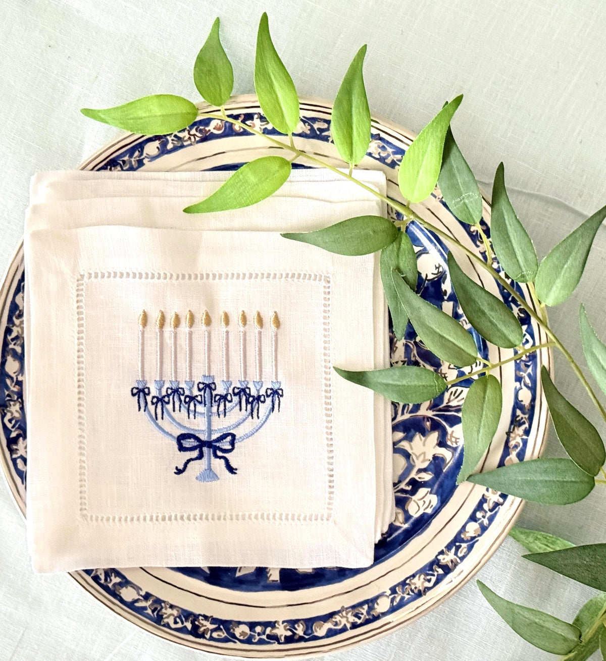 A 6x6 inch 100% linen cocktail napkin, set of 4 embroidered with a light blue Hanukkah Menorah that has dark blue ribbons and gold thread flames set on a Hanukkah plate with a green branch.