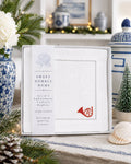 A boxed set of Sweet Humble Home 100% linen cocktail napkins displayed on a tabletop with holiday decor, including pine branches, flowers, a small tree, and blue-and-white ceramic vessels in the background.