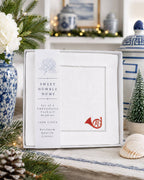 A boxed set of Sweet Humble Home 100% linen cocktail napkins displayed on a tabletop with holiday decor, including pine branches, flowers, a small tree, and blue-and-white ceramic vessels in the background.
