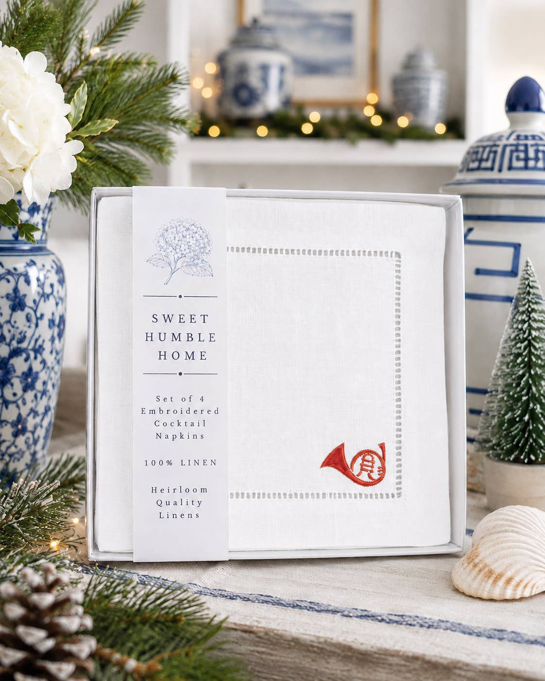 A boxed set of Sweet Humble Home 100% linen cocktail napkins displayed on a tabletop with holiday decor, including pine branches, flowers, a small tree, and blue-and-white ceramic vessels in the background.