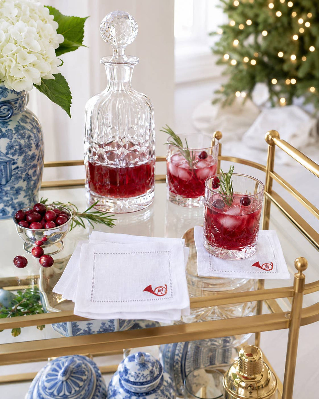 A styled holiday bar scene with a clear cut-glass decanter and two clear rocks glasses filled with red cocktails, set on a gold rolling bar cart. White napkins with a small red embroidered monogram are on the cart, with cranberries, rosemary sprigs, and blue-and-white ceramic vases in the background.