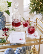 A styled holiday bar scene with a clear cut-glass decanter and two clear rocks glasses filled with red cocktails, set on a gold rolling bar cart. White napkins with a small red embroidered monogram are on the cart, with cranberries, rosemary sprigs, and blue-and-white ceramic vases in the background.