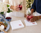 Two people next to a table holding glasses of a dark red cocktail with a rosemary garnish. The scene includes white napkins with a red heart logo, a blue-and-white floral bowl of cranberries, and holiday lights in the background.