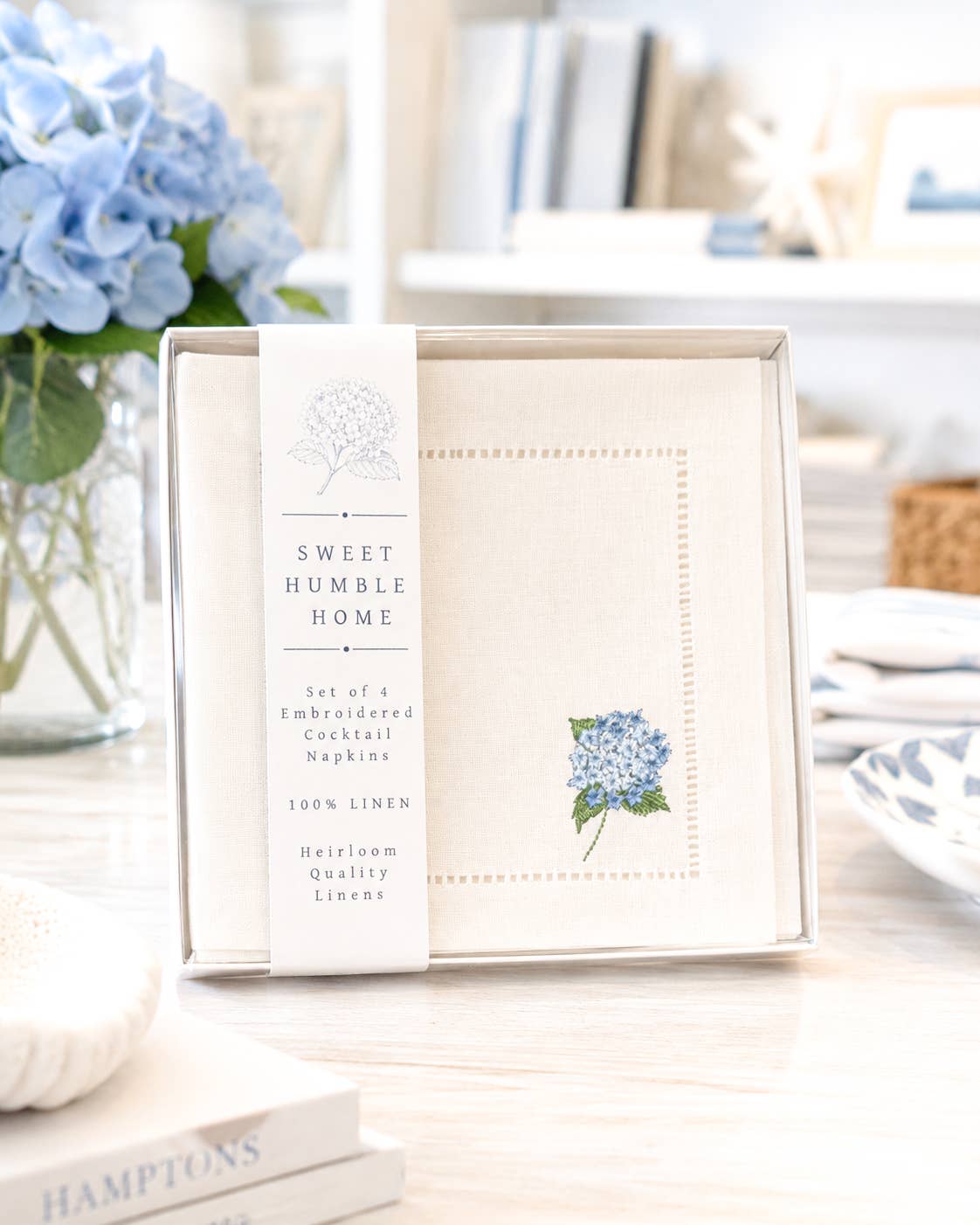 A set of white cocktail napkins in a box ready for gifting. They are 6" square with an embroidered hydrangea in the bottom left corner.
