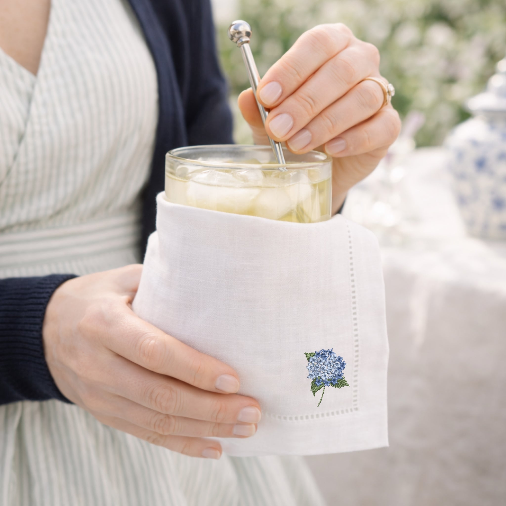 Person holding a glass of lemonade with a white napkin featuring a floral design, outdoors.