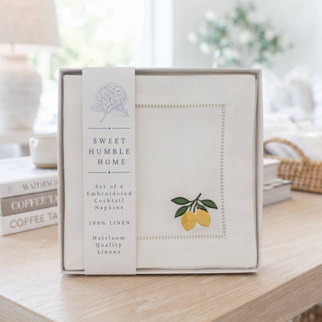 Sweet Humble Home embroidered cocktail napkins set in a gift box displayed on a wooden table in a bright, softly blurred home interior.