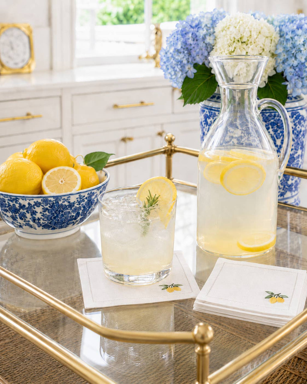 A clear glass carafe filled with lemonade and a matching glass tumbler on a gold-framed bar cart, styled with blue-and-white floral ceramic dishes, lemons, and embroidered white coasters in a bright kitchen setting.