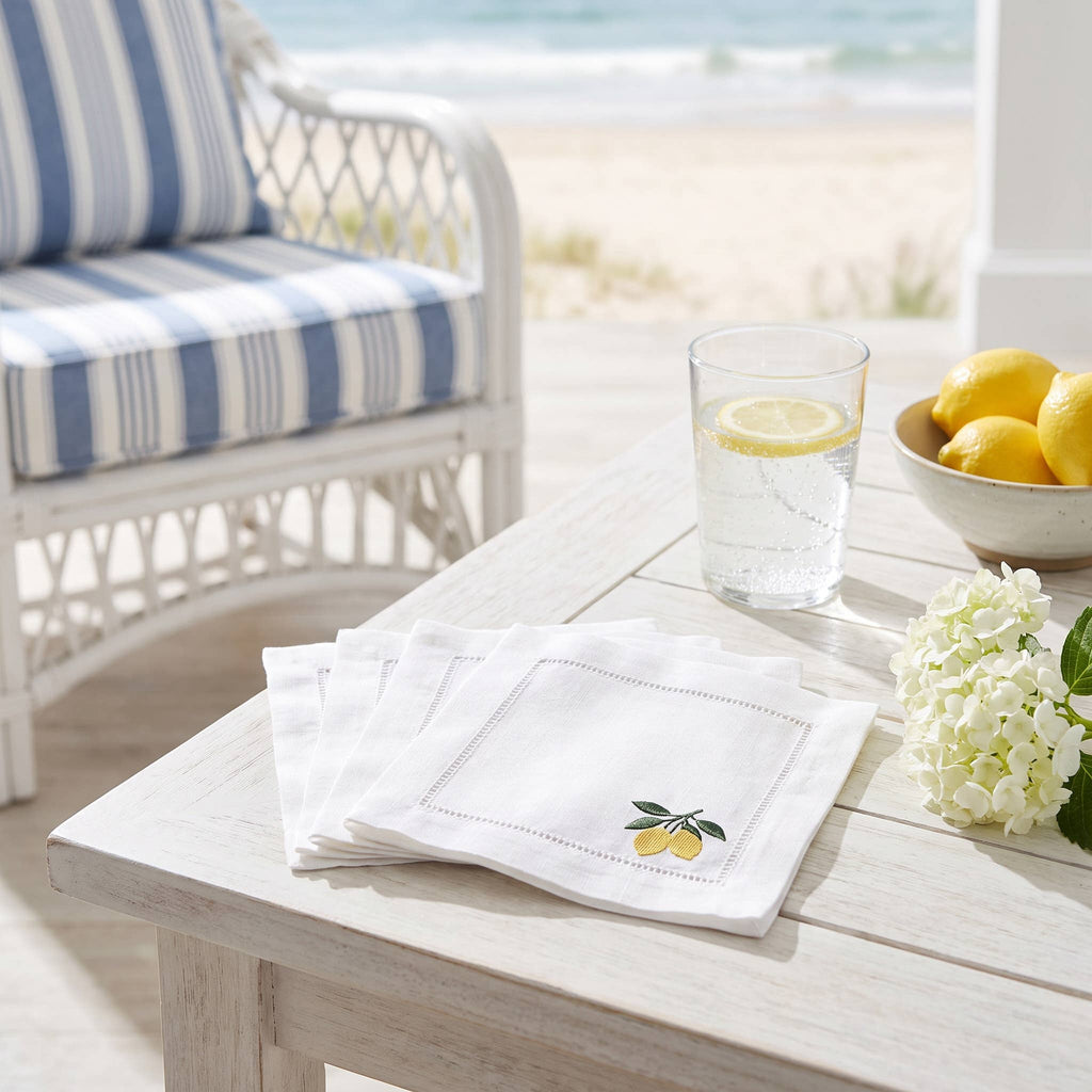 White linen napkins with embroidered lemon motifs arranged on a light wood table in a beachside patio setting, alongside a glass of water with a lemon slice and a bowl of lemons.