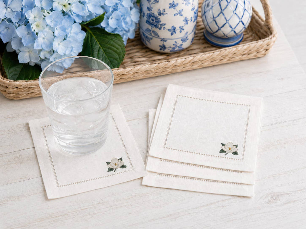 A set of white linen coasters with delicate magnolia embroidery in the corner and a hemstitch border, shown on a light wood table with a glass of water and a stack of coasters. A woven tray with blue flowers and ceramic vases is in the background.