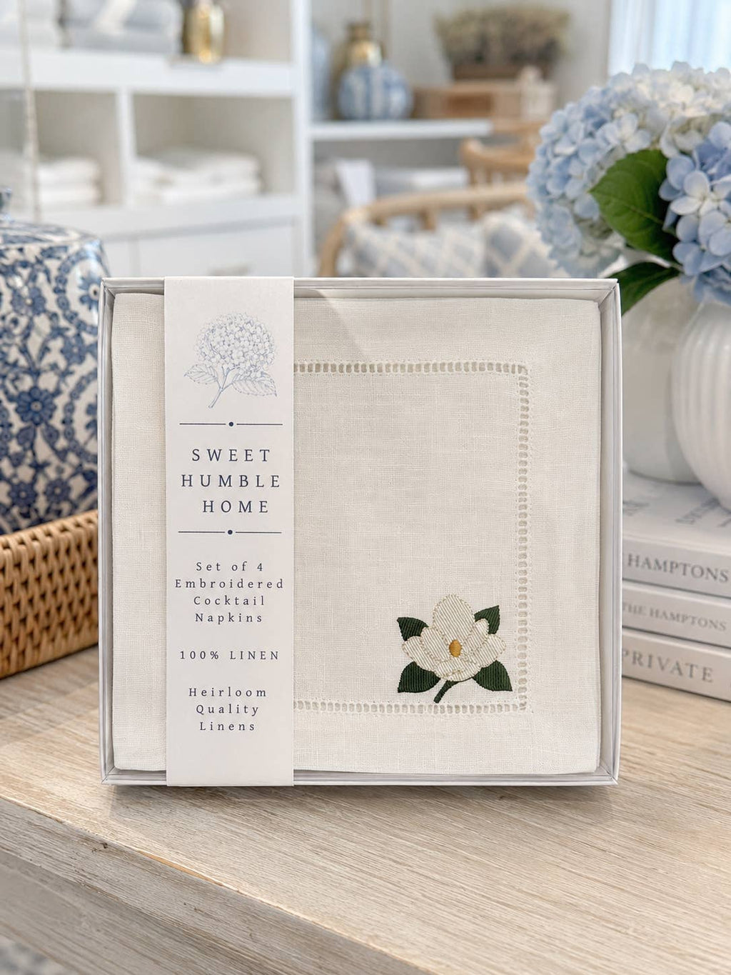 A boxed set of embroidered cocktail napkins on a light wood tabletop, with a floral and home decor styled background. The napkins are white linen with hemstitch borders and white floral magnolia with green leaves.