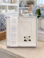 A boxed set of embroidered cocktail napkins on a light wood tabletop, with a floral and home decor styled background. The napkins are white linen with hemstitch borders and white floral magnolia with green leaves.