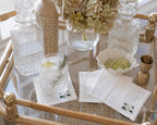A styled bar cart scene with clear glass decanter bottles, a glass of iced drink with rosemary and lime, white embroidered coasters embroidered with a magnolia in the bottom right side above the hemstitch, a small bowl of lime slices, and a brass jigger on a bamboo-and-glass cart.