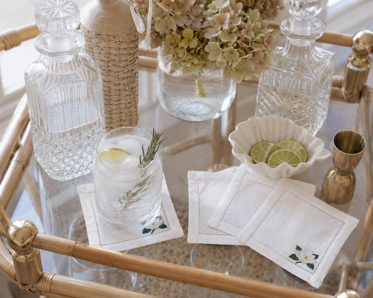 A styled bar cart scene with clear glass decanter bottles, a glass of iced drink with rosemary and lime, white embroidered coasters embroidered with a magnolia in the bottom right side above the hemstitch, a small bowl of lime slices, and a brass jigger on a bamboo-and-glass cart.