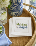 A stack of white embroidered napkins on a woven rattan tray, featuring the text 'Mahjong & Mahgirls' in green and navy blue embroidery. The setting includes a blue and white porcelain vase, a glass of water with a lime slice, and a blue napkin.