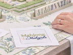 A group of people playing a mahjong game outdoors on a light wood table, surrounded by floral cushions, a glass of rosé wine, and a teacup. There is a linen cocktail napkin that s embroidered with Mahjong & Mahgirls in green and blue